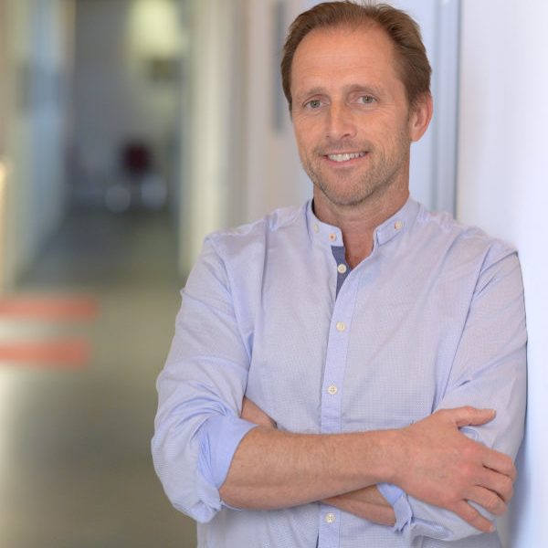 Mann mit lässigem Hemd, lächelnd, in einem modernen Büroflur stehend.