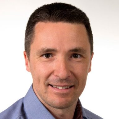 Porträt eines Mannes mit dunklem Haar und einem freundlichen Lächeln. Er trägt ein Hemd.