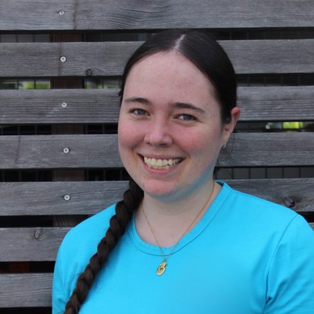 Eine lächelnde Frau mit langem, geflochtenem Haar trägt ein blaues Shirt.