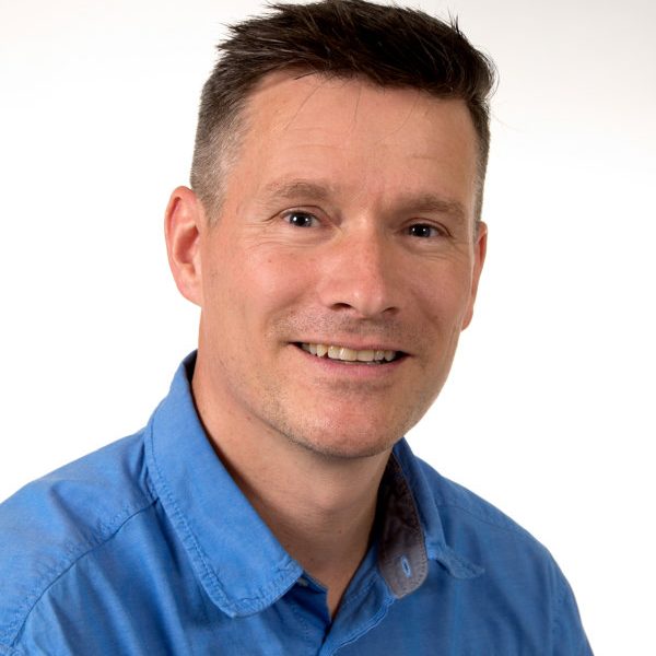 Portrait eines lächelnden Mannes mit kurzem, gestuften Haar, blauem Hemd.