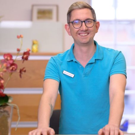 Lächelnder Mann in türkisem Polohemd steht an einem Empfangstresen mit Blumen.