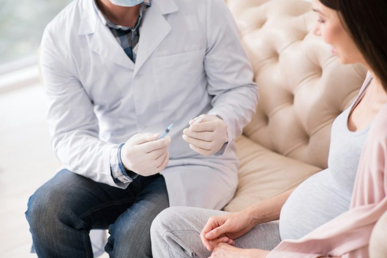 Arzt mit Spritze sitzt neben einer schwangeren Frau auf einer Couch.