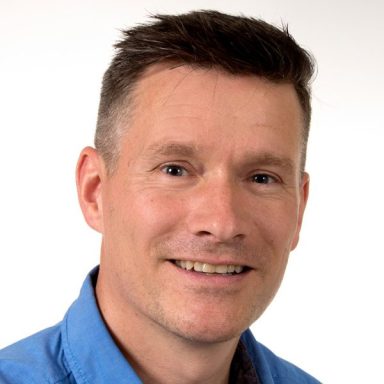 Lächelnder Mann mit kurzem, grauem Haar und blauer Hemd.