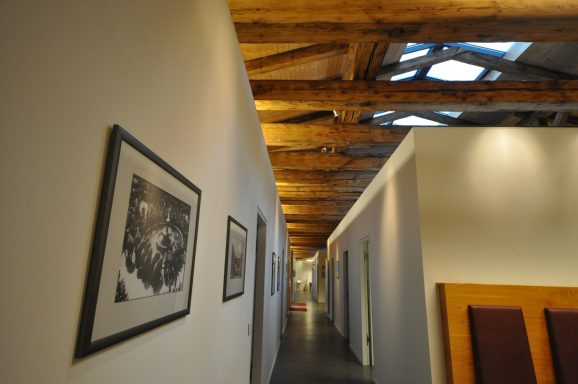 Langer Flur mit hellem Boden, Holzbalken und Schwarz-Weiß-Fotos an den Wänden.