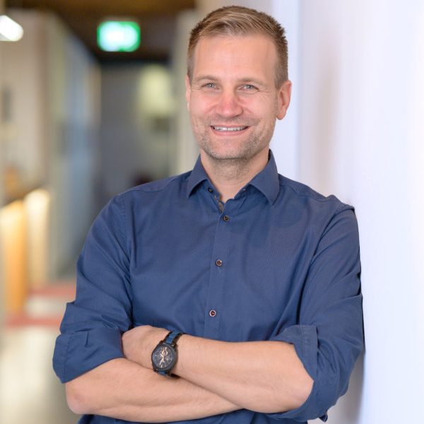 Mann mit kurzen, blonden Haaren, lächelnd, in blauer Hemd, an einer wandstehenden Oberfläche.