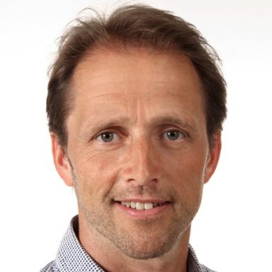 Mann mit kurzen, braunen Haaren und freundlichem Lächeln in kariertem Hemd.