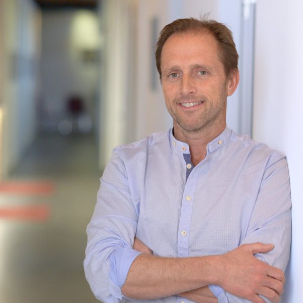 Mann mit verschränkten Armen, lächelnd in einem hellen Flur stehend.