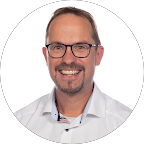 Lächelnder Mann mit Brille, hellen Hemd und kurzen Haaren vor weißem Hintergrund.