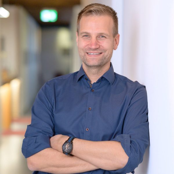 Mann mit kurzem Haar, in blauem Hemd, lächelt und steht mit verschränkten Armen.