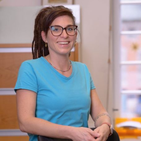 Frau mit Brille und Dreadlocks trägt ein blaues T-Shirt, lächelt freundlich.