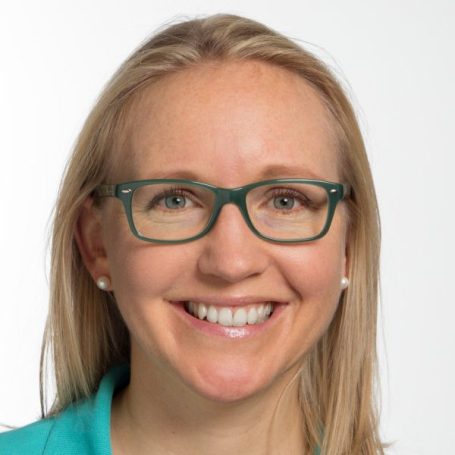 Lächelnde Frau mit blonden Haaren und Brille, trägt ein türkisfarbenes Oberteil.