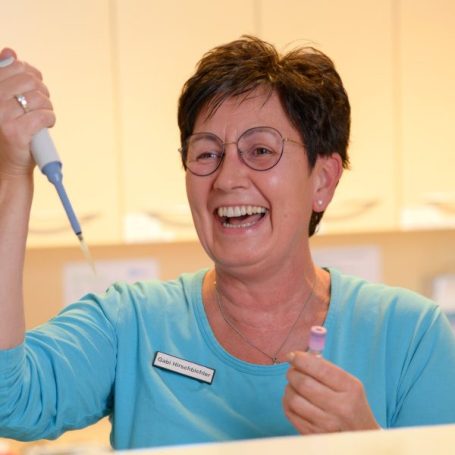 Lächelnde Frau mit Brille hält eine Pipette und eine Probe in der Hand.