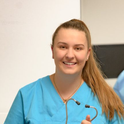 Junge Frau in blauer Arbeitskleidung hält ein Stethoskop, lächelt freundlich.