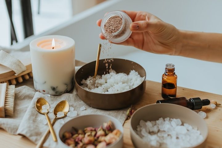 Hände halten ein Glas mit Badesalz neben Schalen, Kerze und ätherischen Ölen.