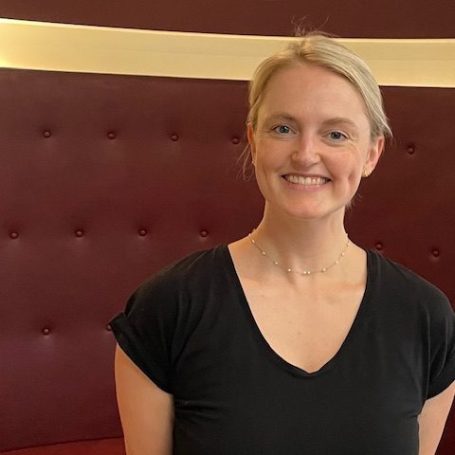 Porträt einer lächelnden Frau mit blonden Haaren vor einem roten Hintergrund.