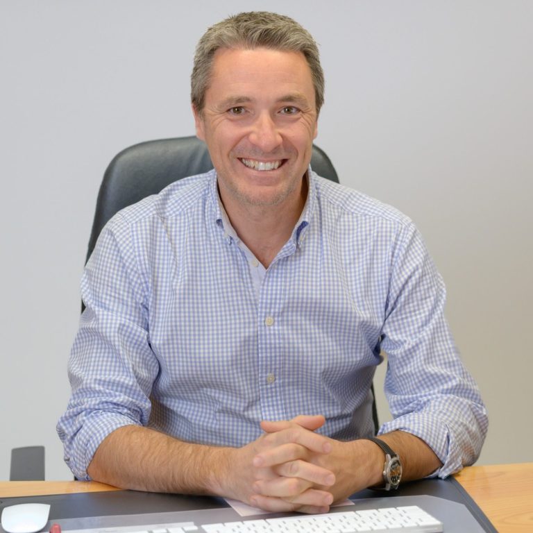 Mann in Hemd mit kariertem Muster, lächelt am Schreibtisch.